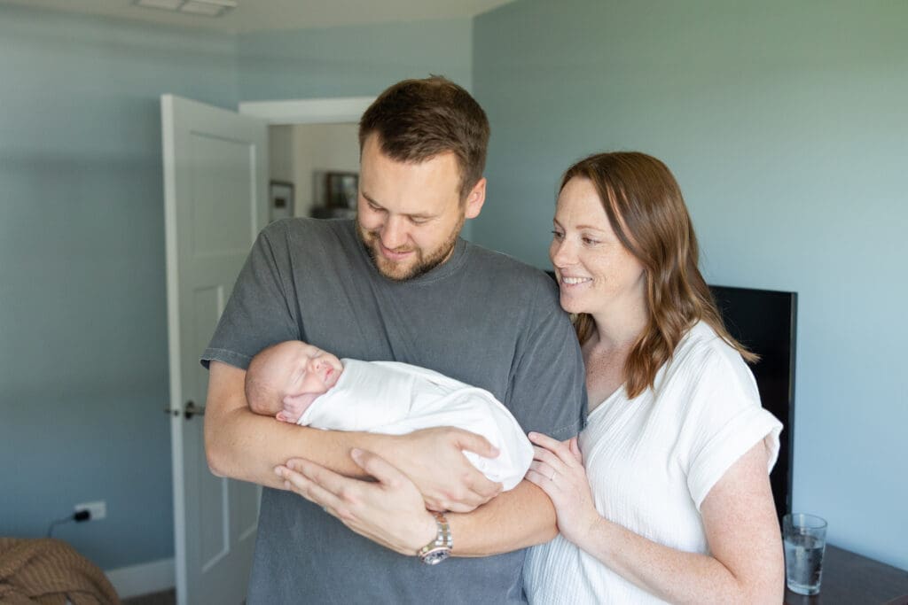 A newborn baby being held by his father while his mother looks on with adoration.