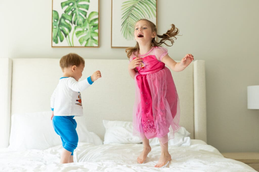 Two young children jumping on the bed with smiles on their faces. 