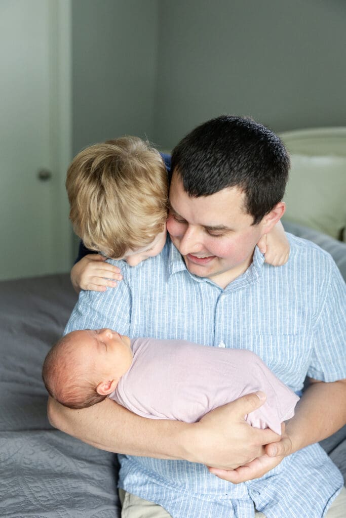 A dad sitting on a bed while his toddler son looks over his shoulder to look at his baby sister resting in daddy's arms.