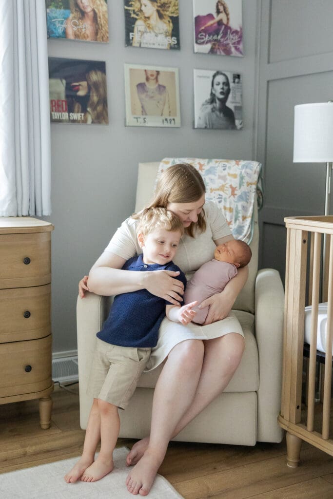 A picture of a mother with her toddler son and baby daughter wrapped in her arms, on a recliner in the baby's nursery.