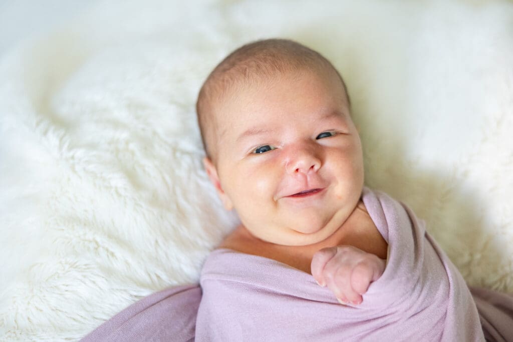 A newborn baby girl is wide-eyed and smiling for the camera.