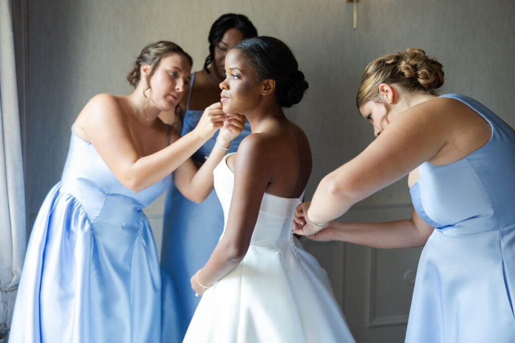 Bridesmaids helping the bride get ready by zipping up her dress and putting earrings on. 
