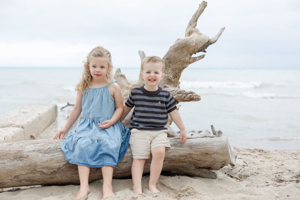 A young brother and sister holding hands while sitting on a log on the beach at Doctors Park. 