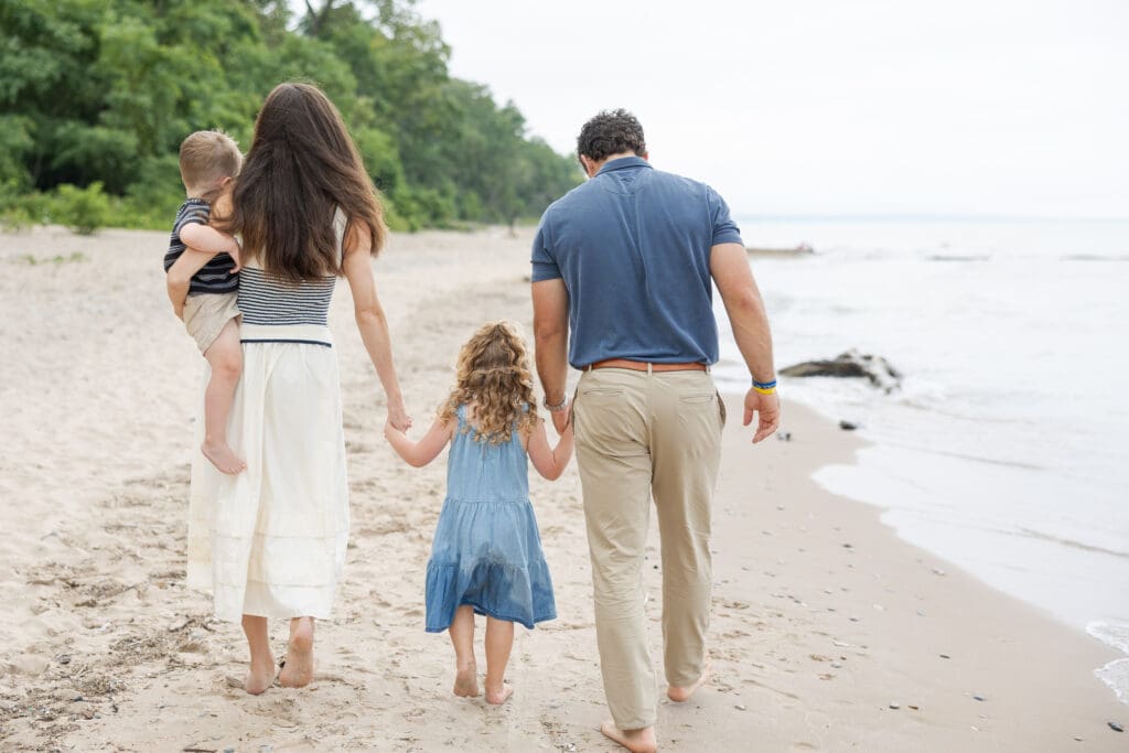 A family of four holding hands and walking away from the camera while on the beach at Doctors Park in Fox Point. 